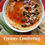 A bowl of creamy, savoury pumpkin hummus topped with seeds, chopped nuts, and a drizzle of sweet chili sauce is served on a red plate with cucumber slices and crackers. Text overlay reads: “Creamy. Comforting. Completely Addictive.”.