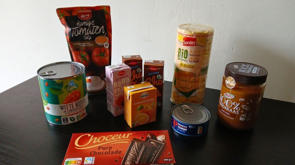 A variety of packaged food items inspired by the Spoonie Survival Guide, including canned beans, tomato sauce, rice cakes, chocolate, peanut butter, juice boxes, canned fish, and instant soup, arranged on a black table against a plain background.
