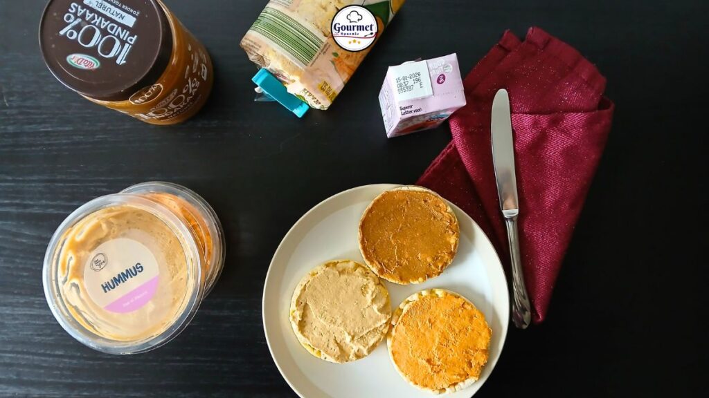A plate with three rice cakes topped with spreads, a knife, and a red napkin on a dark table—perfect zero-prep food. Surrounding items include hummus, peanut butter, packaged cheese slices, and a small carton: true spoonie survival guide essentials.