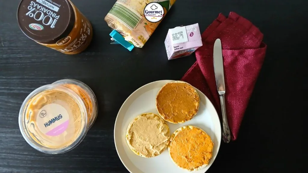 A plate with three rice cakes topped with spreads, a knife, and a red napkin on a dark table—perfect zero-prep food. Surrounding items include hummus, peanut butter, packaged cheese slices, and a small carton: true spoonie survival guide essentials.