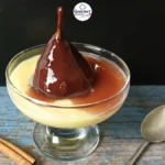 A glass dessert dish with creamy custard and whole Dutch stoofperen (stewed pears) covered in red wine sauce. A cinnamon stick and spoon lie beside it on a rustic blue wooden surface, with a black background and a Gourmet Spoonie logo above.