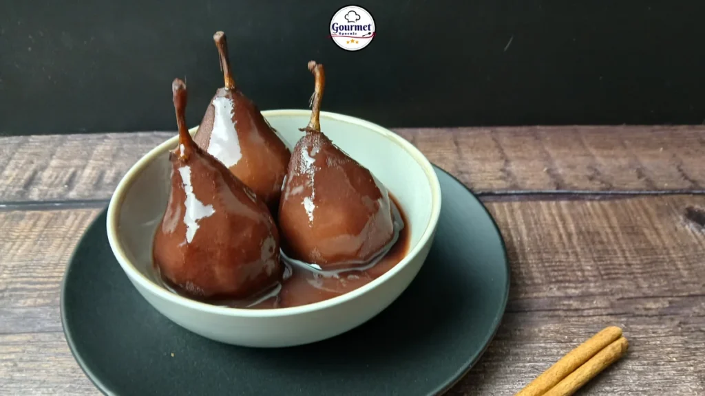 Three stewed pears in spiced wine sit in a light grey bowl on a dark plate. The background is a dark wooden surface, with cinnamon sticks to the side and a Gourmet logo above.