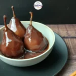 Three stewed pears in spiced wine sit in a light grey bowl on a dark plate. The background is a dark wooden surface, with cinnamon sticks to the side and a Gourmet logo above.