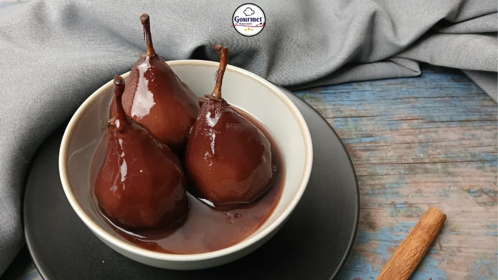 Three Dutch stewed pears coated in glossy dark syrup sit in a light grey bowl on a black plate, with a cinnamon stick and gray cloth nearby on a rustic blue wooden surface.