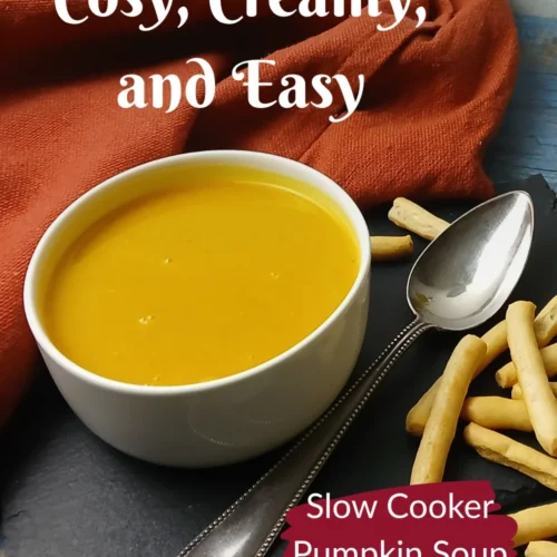 A bowl of creamy slow cooker pumpkin soup with a spoon, surrounded by breadsticks on a slate board. A dish of chopped green onions, a bottle of oil, and an orange napkin are also visible. Text overlay reads: Cosy, creamy, and easy. Slow Cooker Pumpkin Soup.
