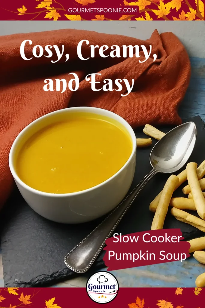 A bowl of creamy slow cooker pumpkin soup with a spoon, surrounded by breadsticks on a slate board. A dish of chopped green onions, a bottle of oil, and an orange napkin are also visible. Text overlay reads: Cosy, creamy, and easy. Slow Cooker Pumpkin Soup.