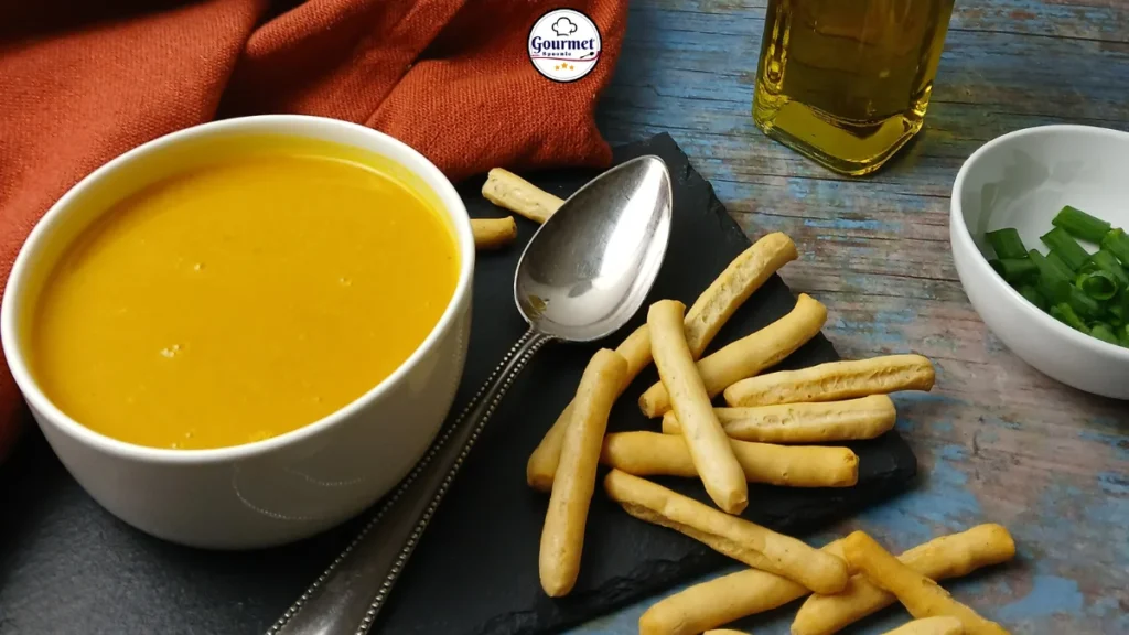 A bowl of creamy slow cooker pumpkin soup with a spoon, surrounded by breadsticks on a slate board. A dish of chopped green onions, a bottle of oil, and an orange napkin are also visible on a rustic blue table.
