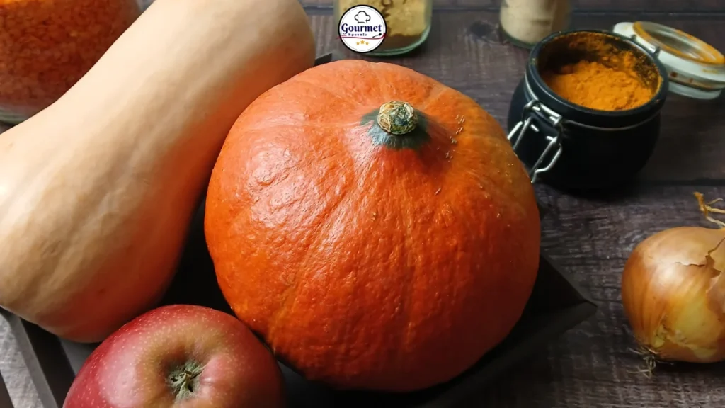 A close-up of red kuri squash, butternut squash, apple, and onion on a dark wooden table, with jars of spices and lentils in the background—perfect ingredients for a cozy slow cooker pumpkin soup.