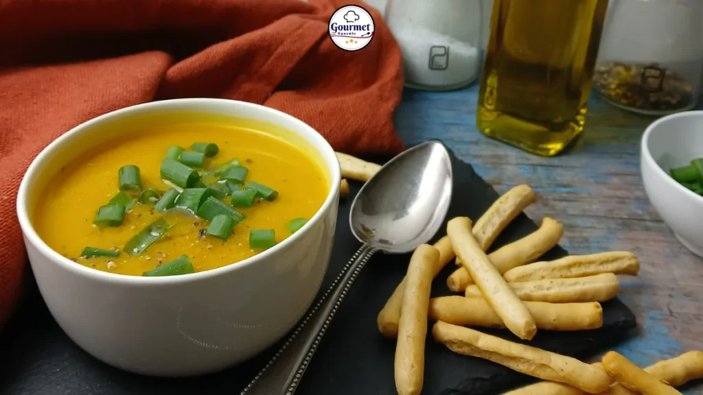 A bowl of creamy pumpkin lentil soup garnished with chopped green onions, served with crispy breadsticks and a spoon on a black slate, with a bottle of olive oil and seasonings in the background.