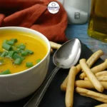 A bowl of creamy pumpkin lentil soup garnished with chopped green onions, served with crispy breadsticks and a spoon on a black slate, with a bottle of olive oil and seasonings in the background.