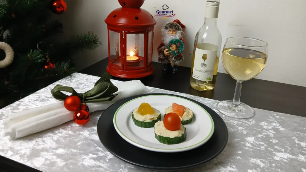A festive table setting with three cucumber canapés, a glass of white wine, a bottle of Chardonnay, a red lantern with a candle, a small Santa figurine, and a white napkin tied with a green ribbon and ornaments.