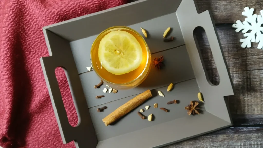 A tray holds a glass of festive drink with lemon, surrounded by whole spices like cinnamon, cardamom, cloves, and star anise—classic gluhwein accents. A red cloth and a white snowflake decoration complete the cosy scene.