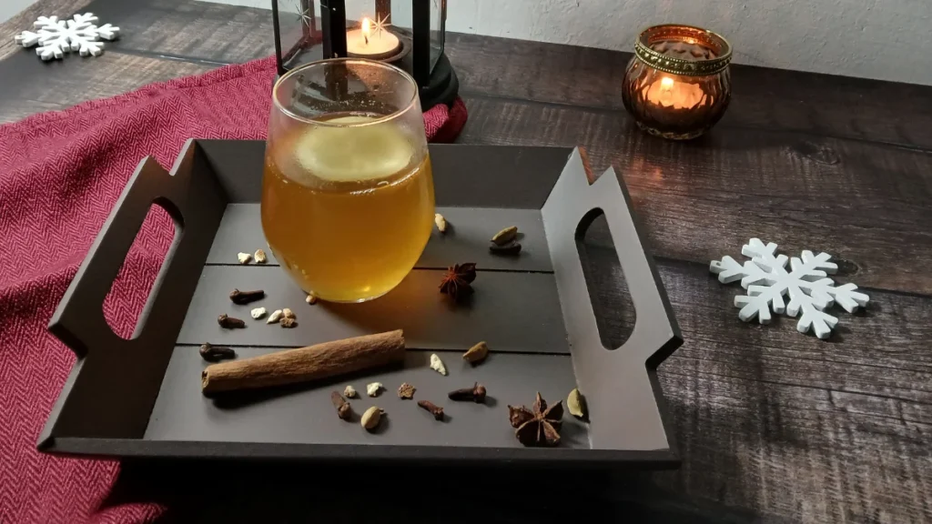 A glass of mulled white wine sits on a wooden tray with cinnamon, star anise, cloves, and cardamom, next to a red cloth. Two lit candles and white snowflake decorations are on a dark wooden table.