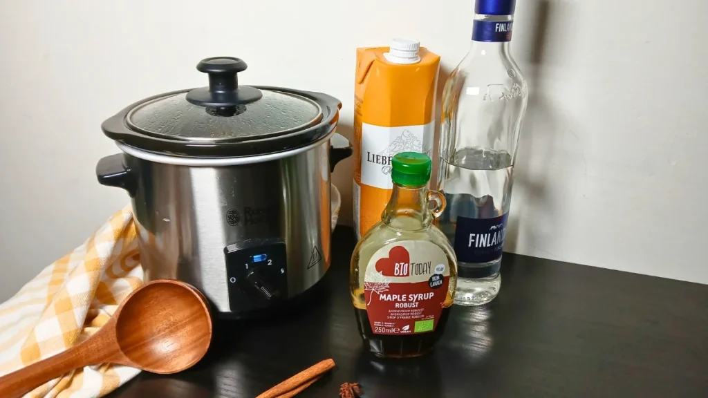A slow cooker, wooden spoon, yellow chequered towel, carton of white wine, bottle of vodka, bottle of maple syrup, and a cinnamon stick are arranged on a dark table—ready for making mulled white wine.