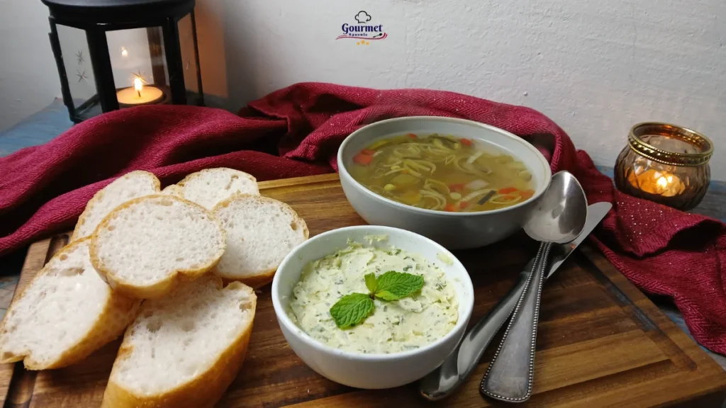 A wooden board with sliced baguette, a bowl of herb butter garnished with mint, and a bowl of Slow Cooker Vegetable Soup. Spoons, a red cloth, and lit candles in the background complete this cozy setting.
