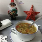 A bowl of vegetable soup with a spoon sits on a table decorated with Christmas ornaments—an inviting scene for an easy Christmas dinner, complete with a napkin with baubles, a lantern, lit candle, Santa figurine, and red star against a white wall.