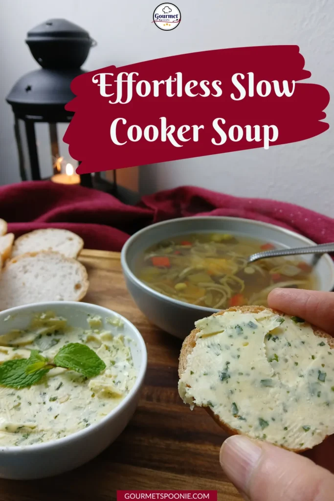 A hand holds a slice of bread with herb butter beside a bowl of soup, bread slices, and a lantern on a wooden table. Text on the image reads, 'Effortless Slow Cooker Soup.'.