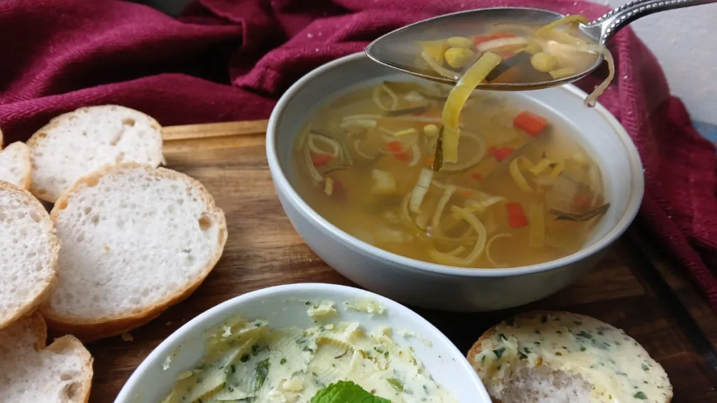 A spoonful of slow cooker soup is held above a bowl on a wooden tray, next to slices of bread and a dish of herbed butter. A red cloth is in the background.