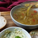 A spoonful of slow cooker soup is held above a bowl on a wooden tray, next to slices of bread and a dish of herbed butter. A red cloth is in the background.