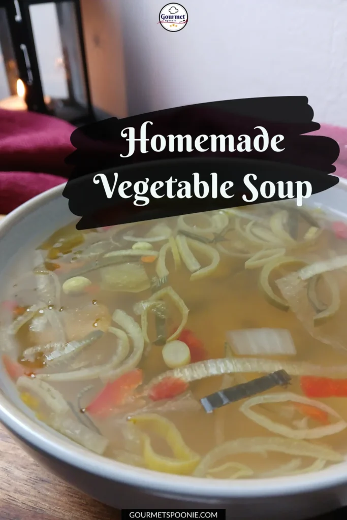 A bowl of clear homemade vegetable soup with sliced onions, carrots, and peas sits on a table. Homemade Vegetable Soup is written above the bowl in white script on a black brushstroke background.