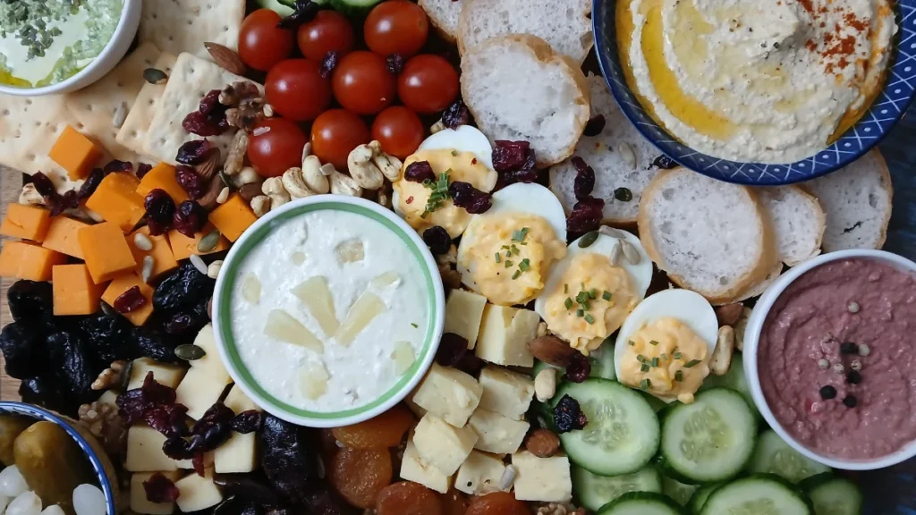 A colourful vegetarian snack board with sliced bread, hummus, devilled eggs, cheeses, nuts, cherry tomatoes, cucumber, crackers, dips, and dried fruits arranged neatly.
