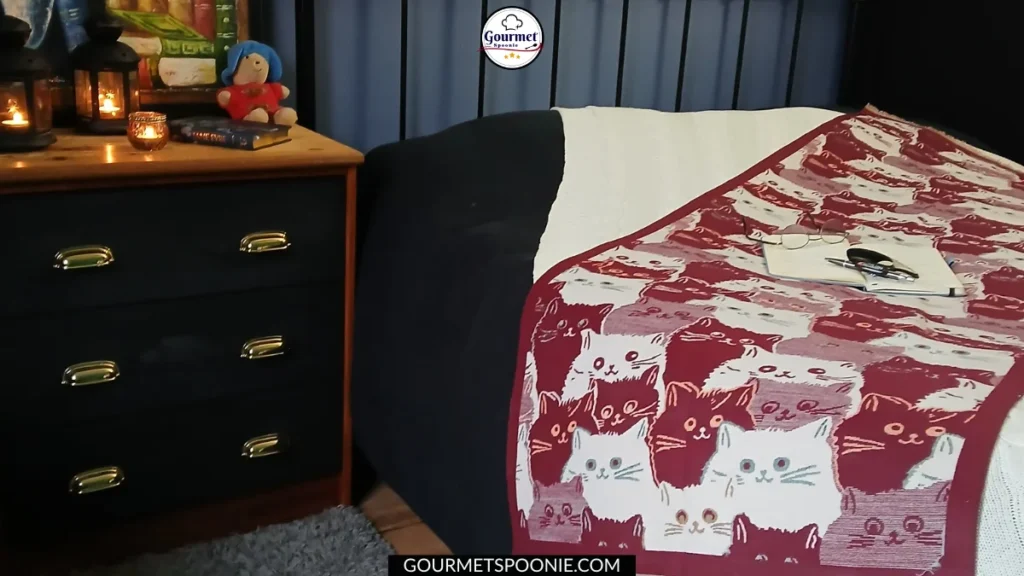 A cosy bedroom, perfect for recovering from a post-holiday burnout, with a bed draped in a red and white cat-patterned blanket. A notebook and glasses rest atop it, while the wooden nightstand holds candles, books, and a Paddington toy.