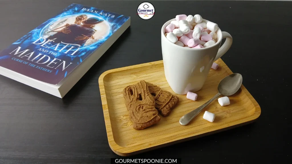 A wooden tray holds two gingerbread biscuits, a silver spoon, and a white mug of hot chocolate with pink and white marshmallows. Next to it rests a fantasy novel titled Death and the Maiden.