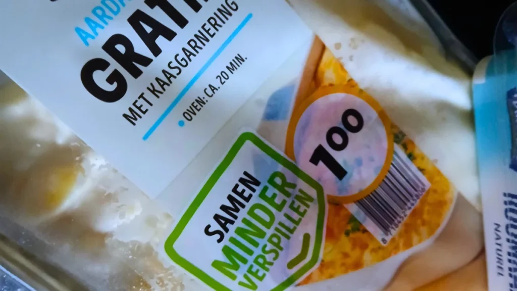 Close-up of a packaged food label in Dutch, showing a dish with cheese topping—ideal spoonie food. The label features oven instructions, a price of 1 euro, and a green sticker: SAMEN MINDER VERSPILLEN (Together less waste).