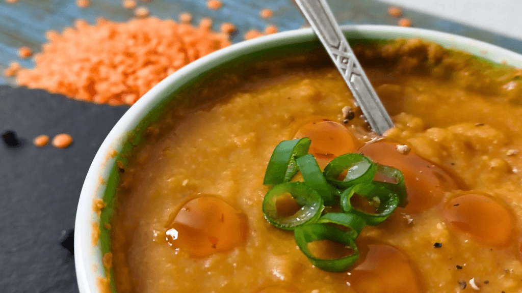 A bowl of comforting lentil soup, thick and orange, is topped with sliced spring onions and a few drops of oil, with a spoon inside. Uncooked red lentils are scattered in the background.