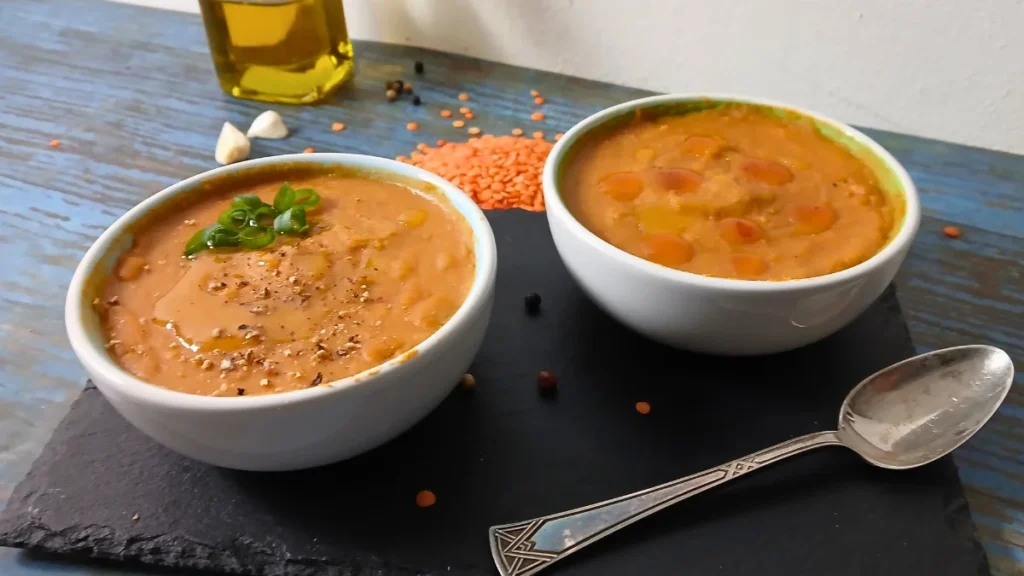 Two white bowls filled with easy lentil soup sit on a dark slate platter. One bowl is garnished with green herbs and pepper, whilst red lentils, garlic, peppercorns, and a bottle of oil appear in the background.