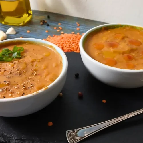 Two white bowls filled with easy lentil soup sit on a dark slate platter. One bowl is garnished with green herbs and pepper, whilst red lentils, garlic, peppercorns, and a bottle of oil appear in the background.