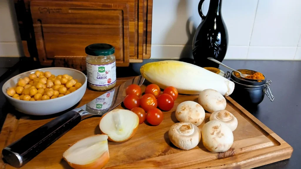 A wooden chopping board with sliced onion, whole mushrooms, cherry tomatoes, Belgian endives, chickpeas, diced garlic, and tikka masala—ingredients perfect for accessible cooking. A large knife and a bottle of olive oil complete this low-effort dinner setup.