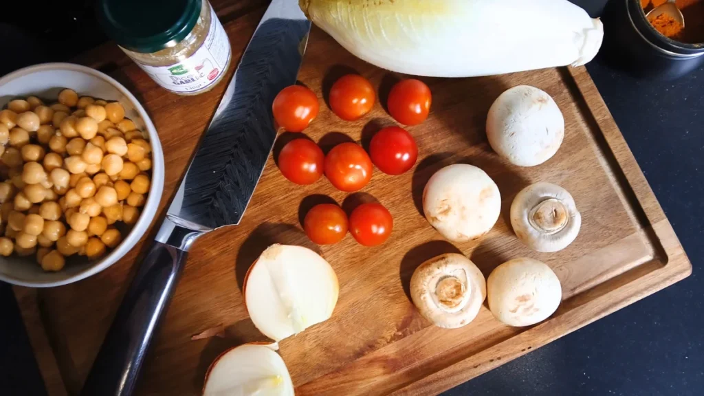 A wooden chopping board with a knife, cherry tomatoes, white mushrooms, half an onion, Belgian chicory, a bowl of chickpeas, a spice jar, and a small container—a perfect setup for an accessible dinner or quick spoonie meal hack on a dark worktop.