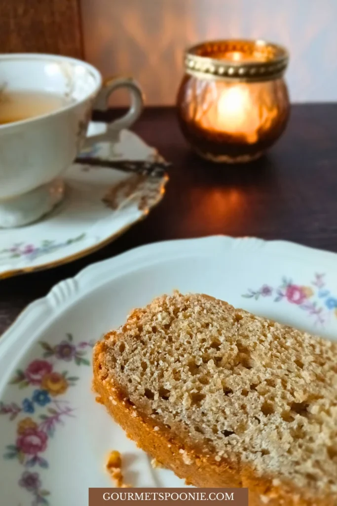 A slice of gluten-free banana bread on a floral plate sits next to a cup of tea and a lit candle on a wooden table.