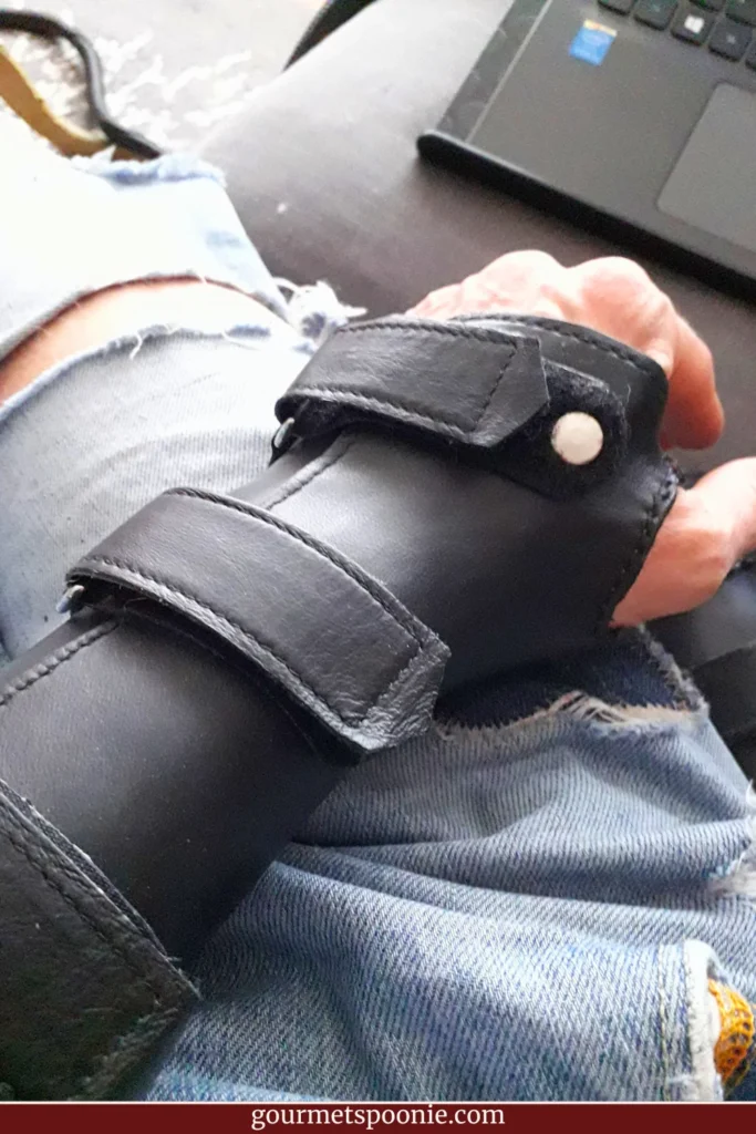 A person with a chronic illness sits next to a laptop, wearing a black leather wrist brace and ripped blue jeans.