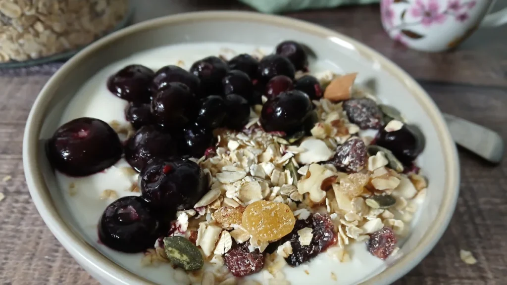 A bowl of yoghurt topped with dark cherries, easy gluten-free muesli, nuts, seeds, and dried fruit sits on a wooden surface.