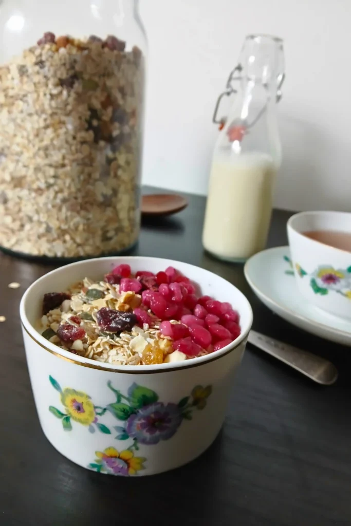A bowl of healthy breakfast muesli topped with pomegranate seeds sits on a dark table next to a jar of muesli, a bottle of non-dairy milk, and a cup of tea.