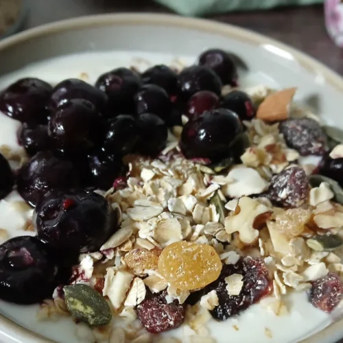 A bowl of yoghurt topped with dark cherries, easy gluten-free muesli, nuts, seeds, and dried fruit sits on a wooden surface.