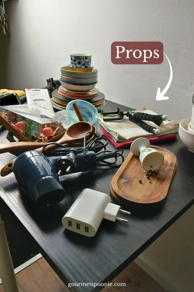 A table cluttered with stacked bowls, wooden utensils, a camera tripod, a hair dryer, a charger, and kitchen items, with a label pointing to Props.