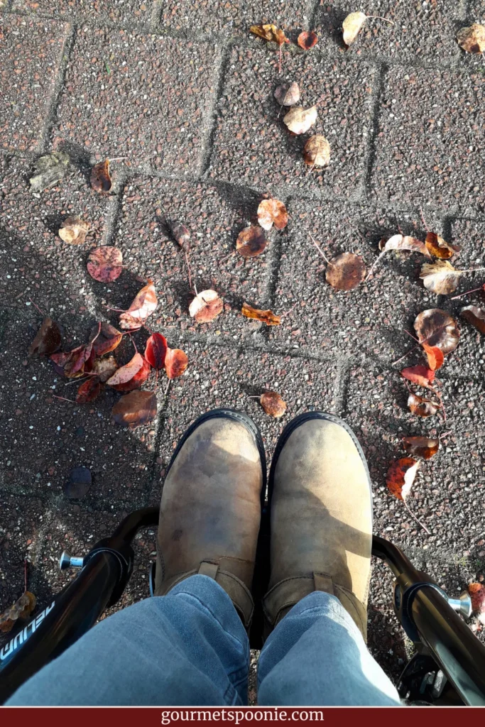 A person embracing disability identity wears tan boots and blue jeans while sitting in a wheelchair on a brick pavement scattered with autumn leaves.