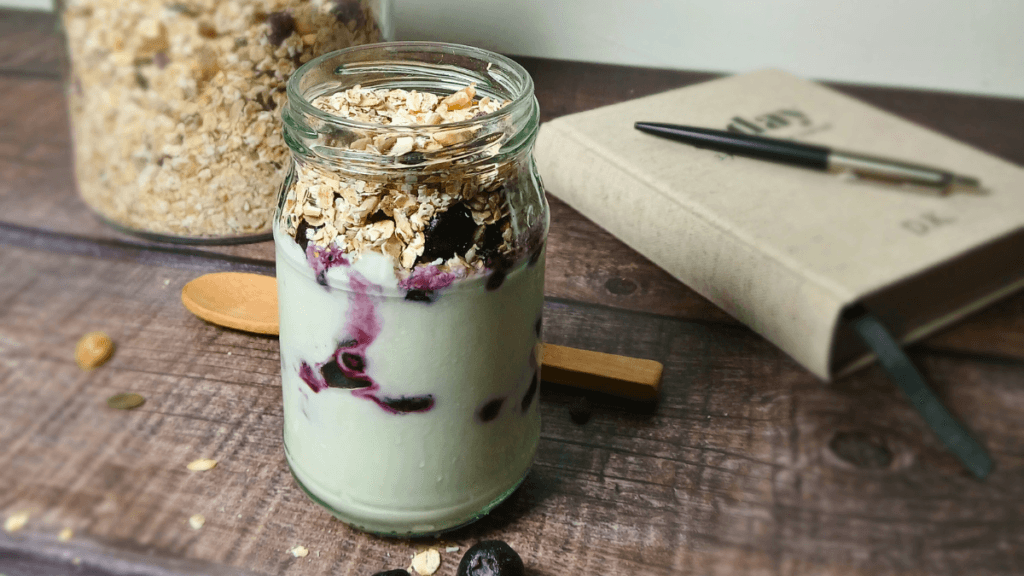 A glass jar filled with a delicious Muesli and Yoghurt Parfait, layered with blueberries and granola, sits on a wooden table next to a notebook, a pen, and a wooden spoon.