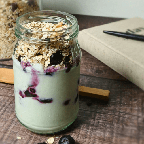 A glass jar filled with a delicious Muesli and Yoghurt Parfait, layered with blueberries and granola, sits on a wooden table next to a notebook, a pen, and a wooden spoon.