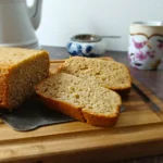 Two slices of banana bread are cut from a loaf on a wooden chopping board, with a teapot, sugar bowl, and floral teacup in the background.
