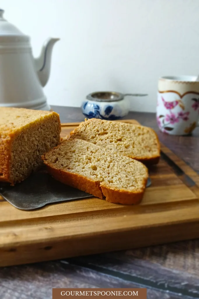Two slices of banana bread are cut from a loaf on a wooden chopping board, with a teapot, sugar bowl, and floral teacup in the background.