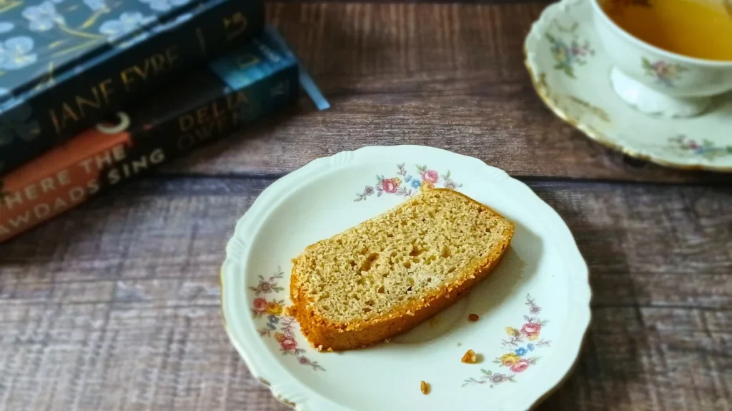 A slice of banana bread on a floral plate sits next to a teacup and saucer, with stacked books in the background on a wooden surface.