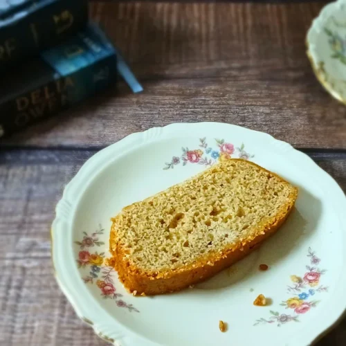 A slice of banana bread on a floral plate sits next to a teacup and saucer, with stacked books in the background on a wooden surface.