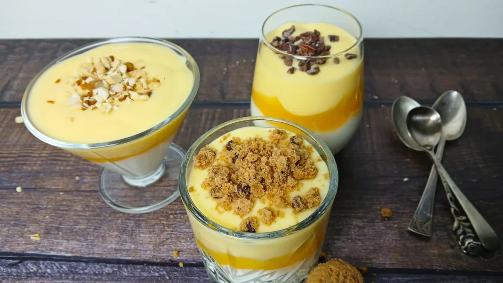 Three glasses of Dutch vlaflip dessert with yoghurt, squash, and Dutch custard vla, each topped with chopped nuts, raisins, or biscuit crumbs, are placed on a wooden surface beside two teaspoons.