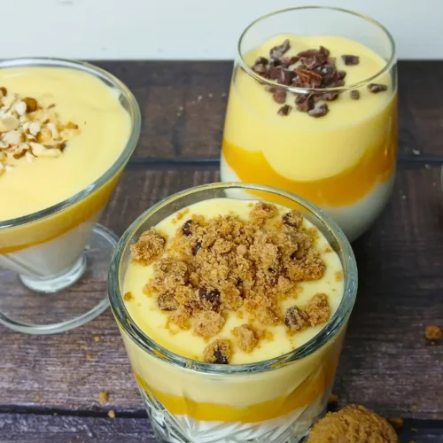 Three glasses of Dutch vlaflip dessert with yoghurt, squash, and Dutch custard vla, each topped with chopped nuts, raisins, or biscuit crumbs, are placed on a wooden surface beside two teaspoons.