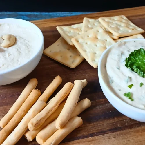 Two bowls of high-protein vegan cream cheese—one topped with cashews, the other with parsley—served alongside breadsticks and square crackers on a wooden board.