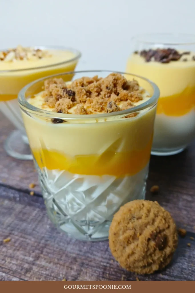 Three layered dessert parfaits in glass cups, featuring white, yellow, and orange layers inspired by a vegan vlaflip recipe, topped with crumbled biscuit and a whole biscuit placed in front.